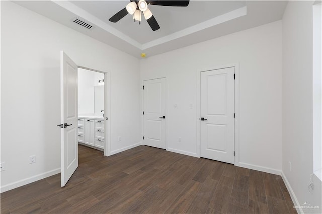 unfurnished bedroom with ceiling fan, a raised ceiling, dark wood-type flooring, and ensuite bath