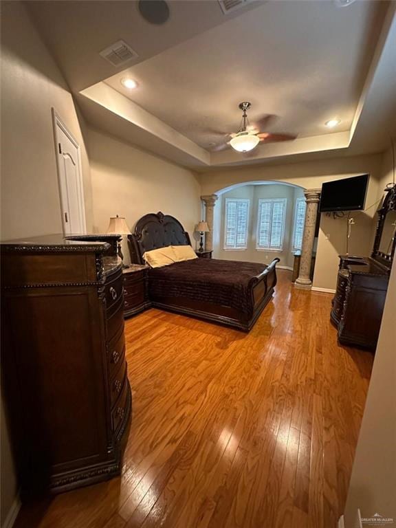 bedroom featuring a raised ceiling, light hardwood / wood-style floors, ornate columns, and ceiling fan