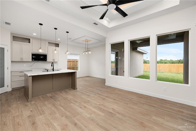 kitchen with light wood-type flooring, a tray ceiling, ceiling fan, decorative light fixtures, and an island with sink