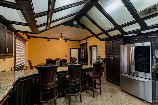 kitchen featuring a breakfast bar, ceiling fan, stainless steel fridge, light stone counters, and dark brown cabinetry