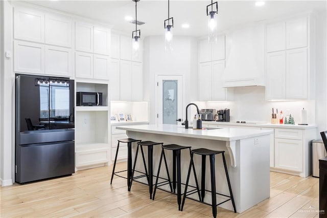 kitchen with custom exhaust hood, sink, black appliances, white cabinets, and an island with sink