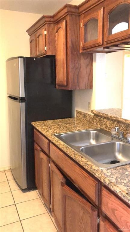 kitchen featuring glass insert cabinets, light tile patterned floors, freestanding refrigerator, brown cabinetry, and a sink