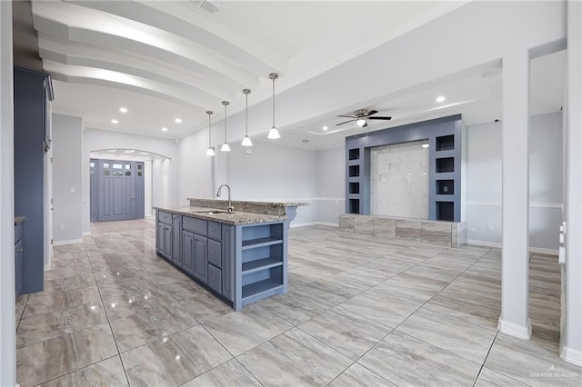 kitchen with light stone countertops, ceiling fan, a kitchen island with sink, sink, and decorative light fixtures