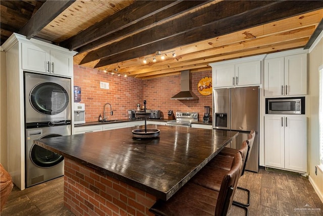 kitchen with dark wood-style flooring, dark countertops, wall chimney exhaust hood, appliances with stainless steel finishes, and stacked washer and clothes dryer