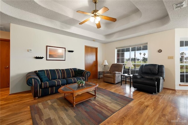 living room with ceiling fan, light wood-type flooring, a textured ceiling, and a tray ceiling