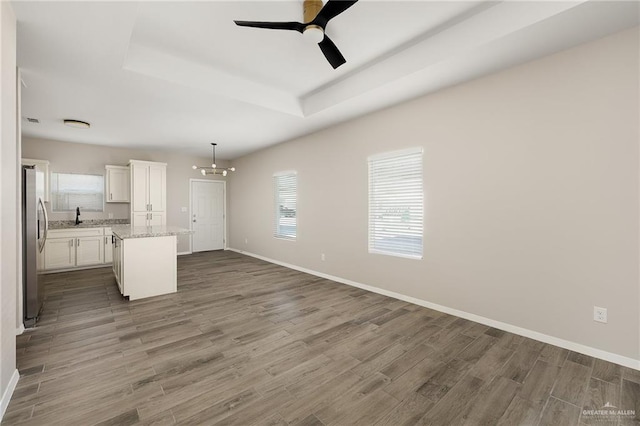 unfurnished living room with a raised ceiling, a sink, wood finished floors, baseboards, and ceiling fan with notable chandelier