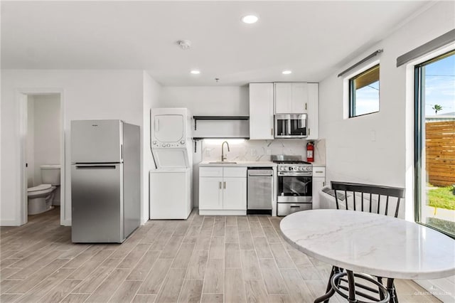 kitchen with tasteful backsplash, sink, white cabinetry, stacked washing maching and dryer, and stainless steel appliances