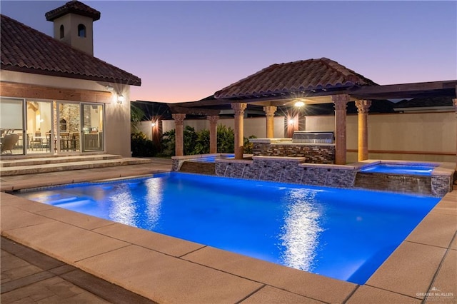 pool at dusk featuring a patio area, a pool with connected hot tub, fence, and an outdoor kitchen