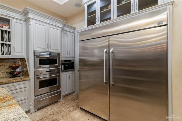 kitchen featuring appliances with stainless steel finishes, light stone countertops, a warming drawer, tasteful backsplash, and glass insert cabinets