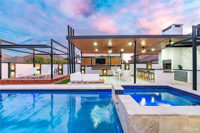 pool at dusk featuring a patio, an in ground hot tub, a bar, pool water feature, and exterior kitchen