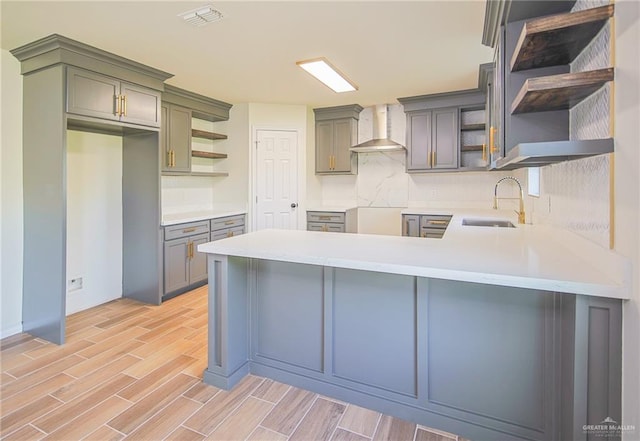 kitchen with wall chimney range hood, sink, light wood-type flooring, tasteful backsplash, and kitchen peninsula