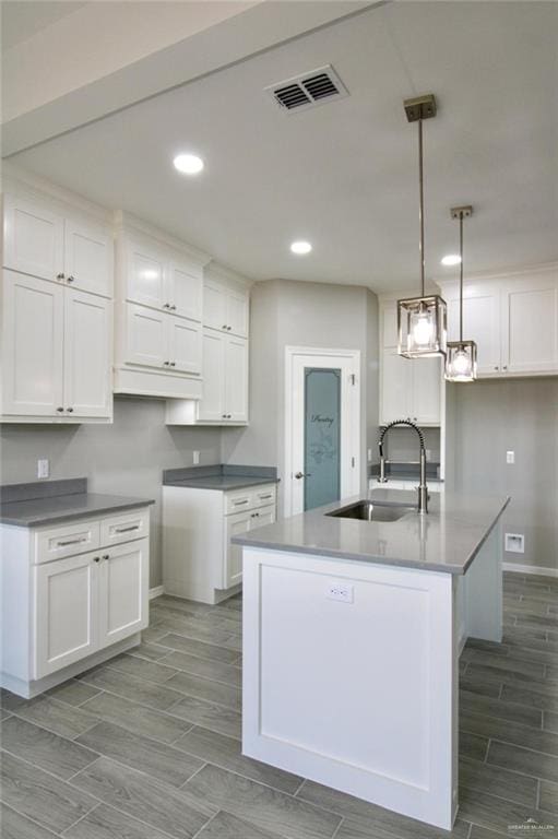 kitchen featuring visible vents, hanging light fixtures, a kitchen island with sink, a sink, and white cabinetry