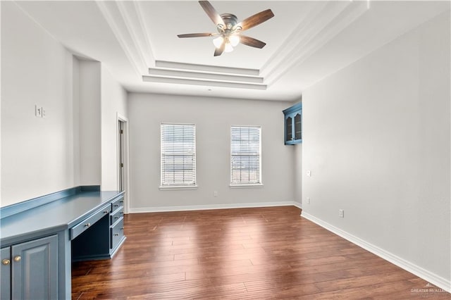 unfurnished living room with a raised ceiling, ceiling fan, built in desk, and dark wood-type flooring