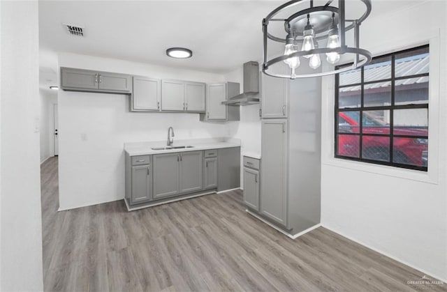 kitchen with gray cabinetry, a notable chandelier, light wood-type flooring, wall chimney range hood, and sink