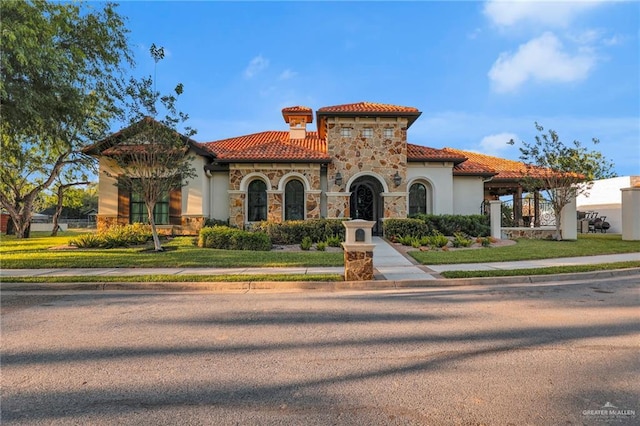 mediterranean / spanish home featuring a tile roof, stone siding, a front lawn, and stucco siding