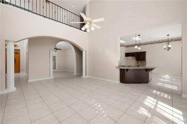 unfurnished living room with sink, a high ceiling, decorative columns, light tile patterned floors, and ceiling fan with notable chandelier