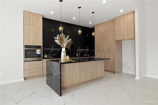 kitchen featuring stainless steel oven, a kitchen island with sink, sink, light brown cabinetry, and decorative light fixtures