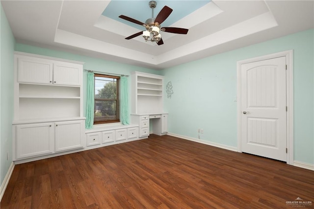 unfurnished bedroom featuring ceiling fan, a tray ceiling, dark hardwood / wood-style flooring, and built in desk