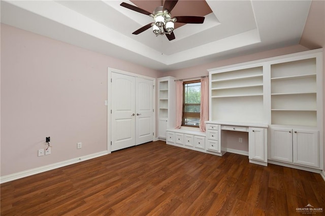 interior space with ceiling fan, a tray ceiling, built in desk, and dark wood-type flooring