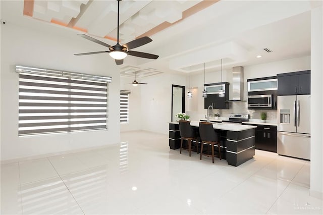 kitchen featuring wall chimney range hood, a kitchen island with sink, hanging light fixtures, stainless steel appliances, and a kitchen bar