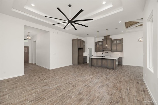 kitchen with ceiling fan, a raised ceiling, light hardwood / wood-style flooring, decorative light fixtures, and a kitchen island with sink