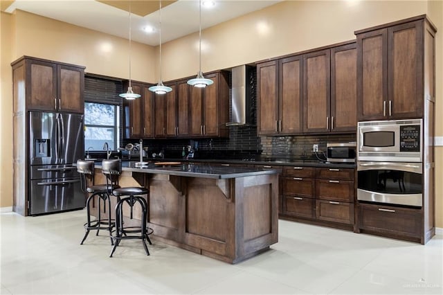 kitchen featuring appliances with stainless steel finishes, a kitchen breakfast bar, an island with sink, pendant lighting, and wall chimney range hood