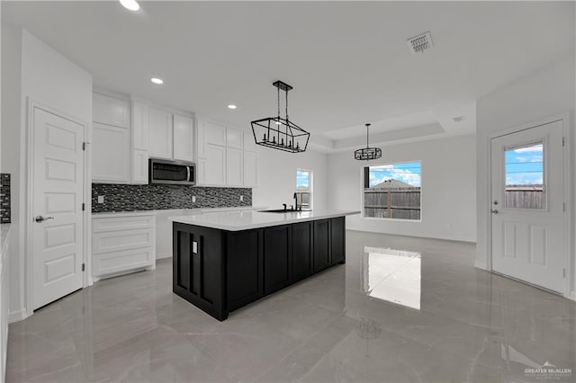 kitchen featuring a raised ceiling, decorative light fixtures, decorative backsplash, a center island with sink, and white cabinets