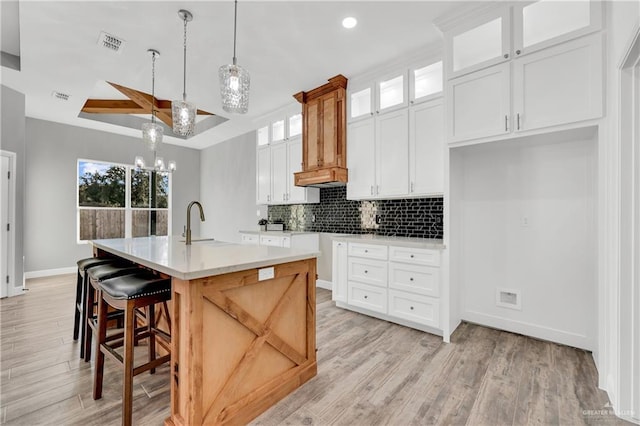 kitchen with white cabinetry, backsplash, a kitchen island with sink, hanging light fixtures, and light wood-type flooring