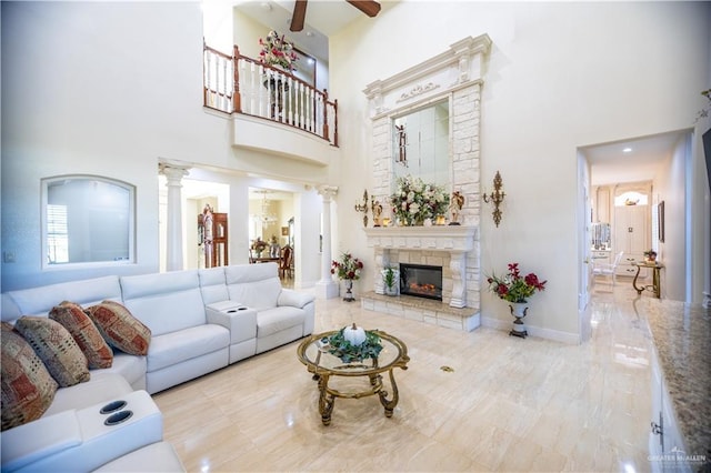 living area featuring a towering ceiling, a ceiling fan, ornate columns, and a stone fireplace