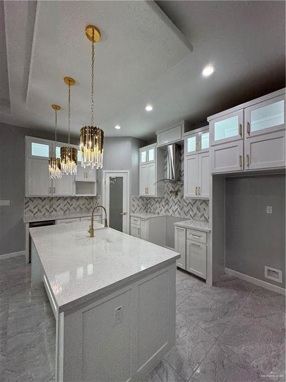 kitchen with pendant lighting, wall chimney range hood, sink, an island with sink, and white cabinets