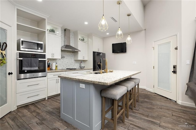 kitchen featuring white cabinets, wall chimney range hood, black appliances, an island with sink, and decorative light fixtures