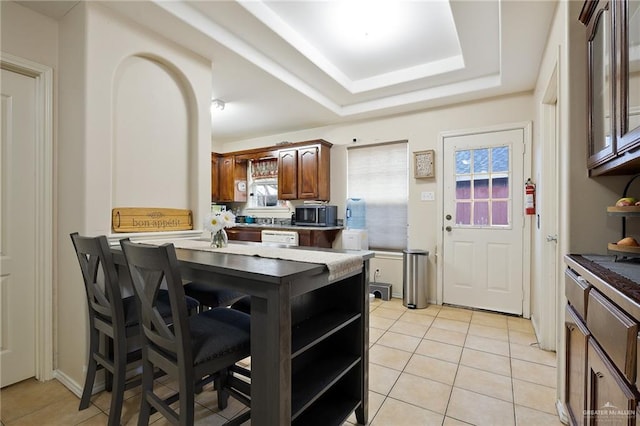 kitchen with a breakfast bar, a tray ceiling, light tile patterned floors, dark countertops, and white dishwasher