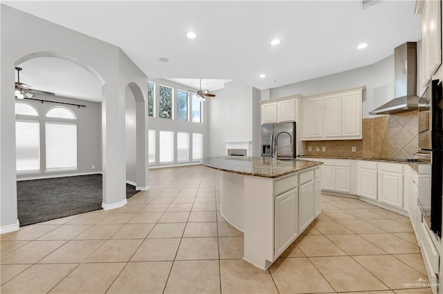 kitchen with a kitchen island with sink, wall chimney range hood, ceiling fan, stone countertops, and light tile patterned flooring