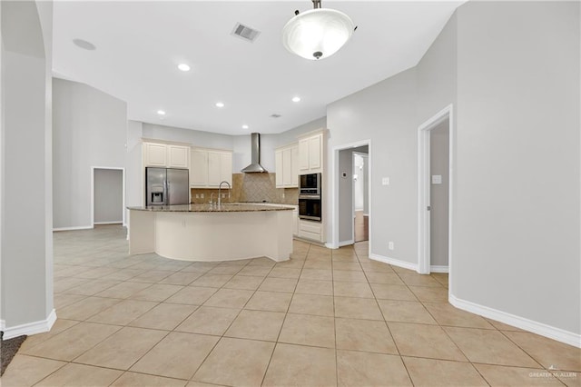 kitchen with wall chimney range hood, dark stone counters, a kitchen island with sink, light tile patterned flooring, and appliances with stainless steel finishes