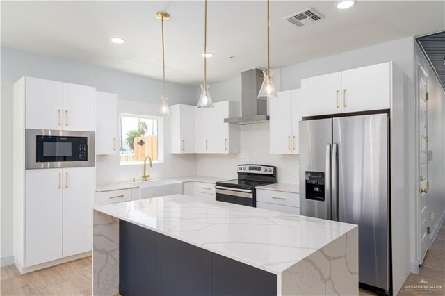 kitchen featuring wall chimney range hood, a kitchen island, pendant lighting, white cabinets, and appliances with stainless steel finishes