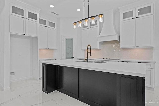 kitchen featuring a center island with sink, white cabinets, glass insert cabinets, custom exhaust hood, and light countertops