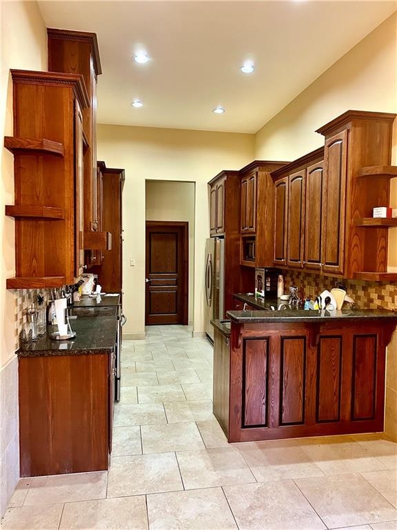 kitchen featuring stove, dark stone counters, decorative backsplash, kitchen peninsula, and stainless steel refrigerator