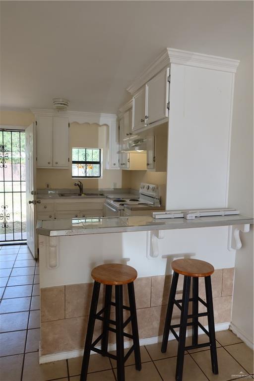 116 N Kansas Ave, Weslaco, TX 78596 - kitchen featuring white electric stove, light tile patterned flooring, a breakfast bar area, light countertops, and white cabinetry