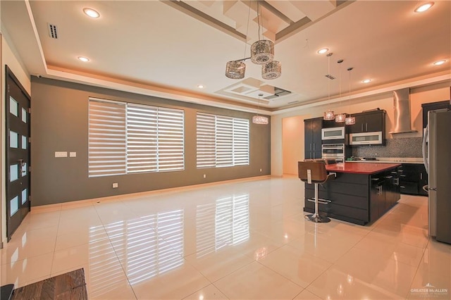 kitchen featuring stainless steel appliances, a raised ceiling, a kitchen island with sink, and wall chimney range hood