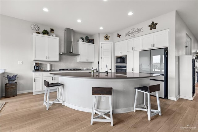 kitchen with wall chimney range hood, white cabinetry, a kitchen breakfast bar, stainless steel appliances, and a center island with sink