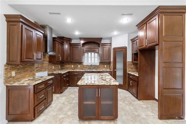 kitchen with light stone countertops, backsplash, wall chimney exhaust hood, sink, and a center island