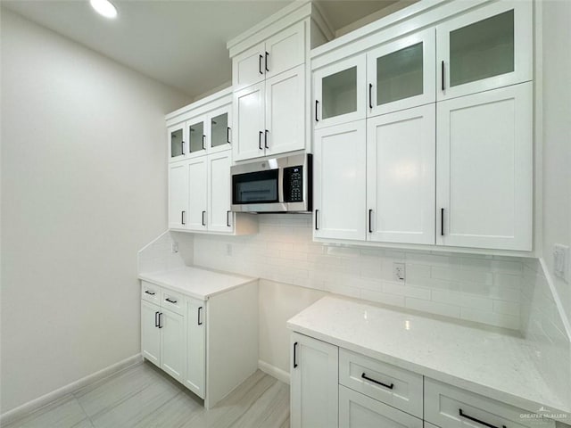 kitchen featuring light stone countertops, glass insert cabinets, white cabinetry, stainless steel microwave, and backsplash