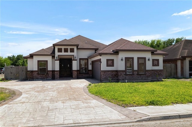 prairie-style home featuring a front yard, an attached garage, fence, stucco siding, and decorative driveway