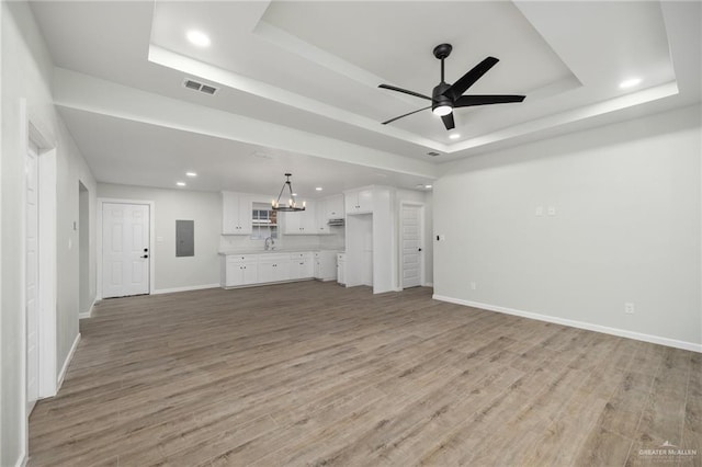 unfurnished living room with ceiling fan with notable chandelier, a tray ceiling, and light hardwood / wood-style floors