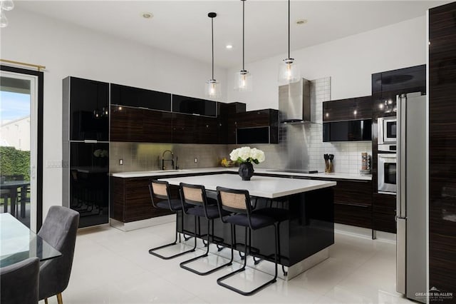 kitchen with wall chimney exhaust hood, appliances with stainless steel finishes, a kitchen island, pendant lighting, and backsplash