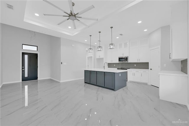 kitchen featuring tasteful backsplash, decorative light fixtures, a raised ceiling, an island with sink, and white cabinets