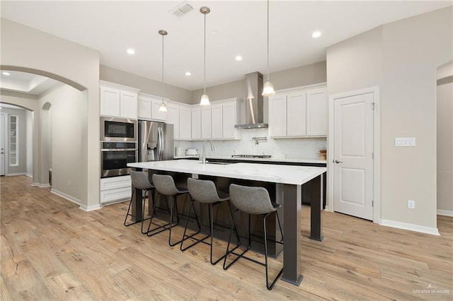 kitchen featuring wall chimney range hood, appliances with stainless steel finishes, an island with sink, white cabinets, and decorative backsplash