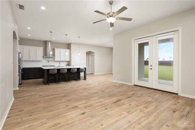 kitchen featuring wall chimney range hood, light hardwood / wood-style flooring, a kitchen island with sink, hanging light fixtures, and white cabinets