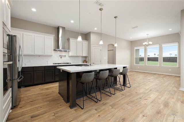 kitchen with white cabinetry, decorative light fixtures, wall chimney range hood, a kitchen island with sink, and backsplash