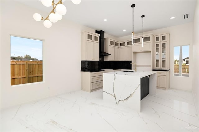 kitchen featuring backsplash, wall chimney range hood, light stone countertops, an island with sink, and decorative light fixtures